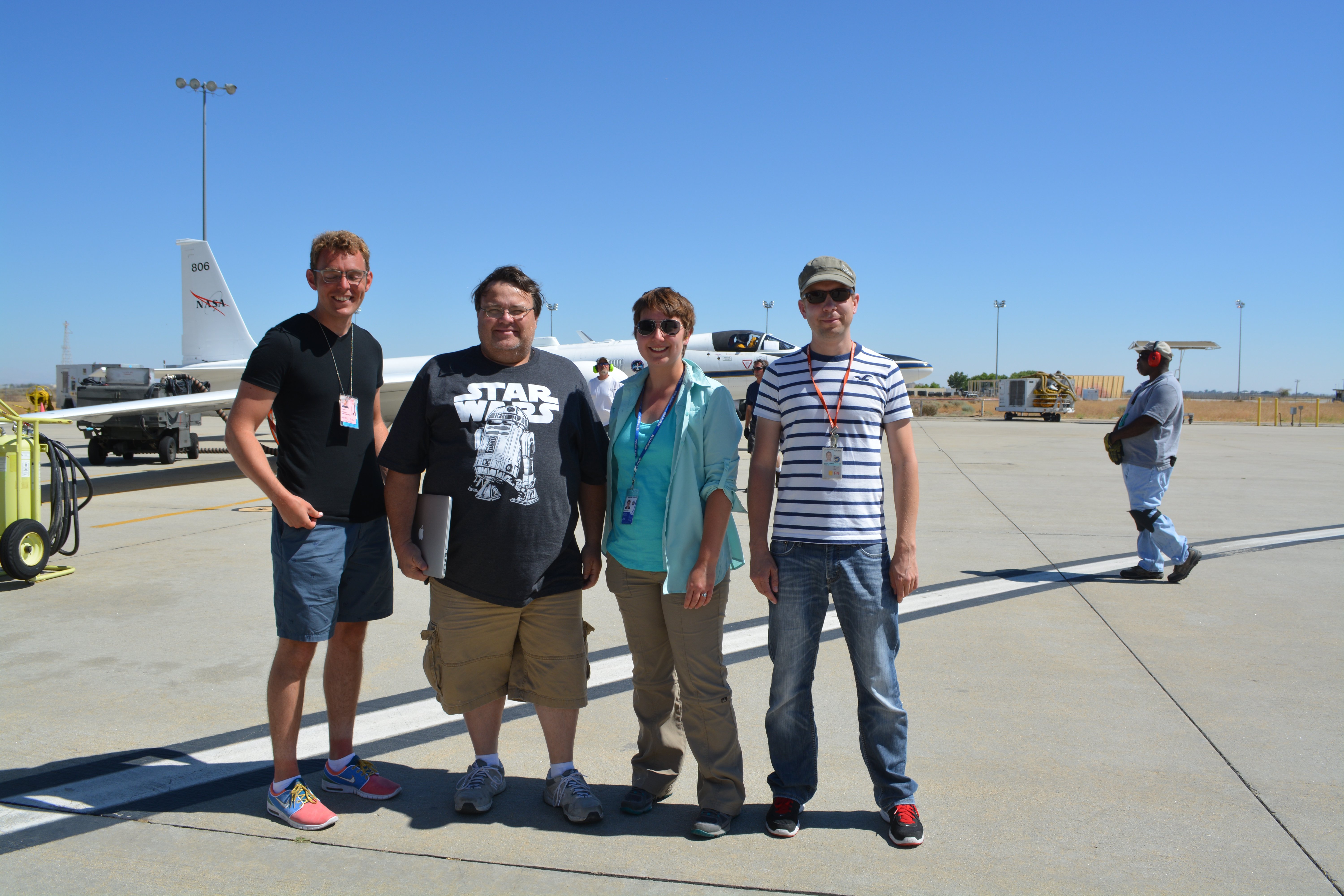An image of four people standing on the apron outside a hangar at Armstrong Flight Research Center. In the background are several other people wearing ear protection and the NASA ER-2 aircraft parked on the apron with the pilot in a flight suit and helmet visible in the cockpit. It is a warm sunny day with a completely blue sky.