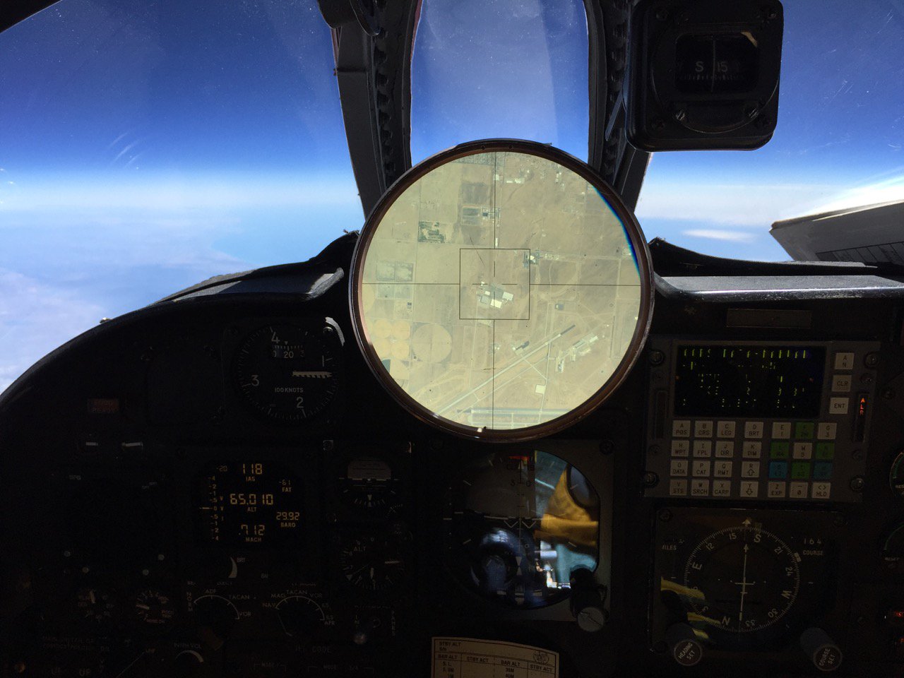 An image of an airplane's cockpit, with various instrument panels visible. The view out of the front of the airplane shows a very deep blue sky above and indistinct looking land below. In the middle of the dash is a large circular instrument showing a much-enlarged view of the ground below the plane, which shows several fields, houses, and runways, and in the center of the crosshairs on the instrument, a few rectangular buildings and parking lots.