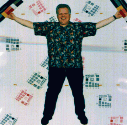 This image was taken using a MISR camera. This poster is of Larry Hovland, the instrument engineer for the project. He holds color tablets, and is surrounded by numerous targets, each a cascade of bar targets decreasing in size. These targets allow us to verify the digital data stream, and our ability to reconstruct an image from packets of data transmitted by the instrument. More sophisticated tests were used to characterize the image quality of each camera. The original poster is 4 x 6 ft in size, and was hung from the ceiling of the high-bay, where MISR was built.Click here to see the laboratory set-up.